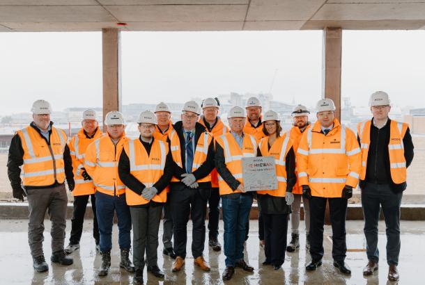 People gathered around two people holding a signed metal plaque to celebrate a construction topping out