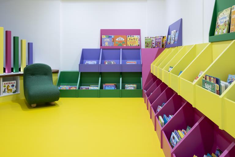A colourfully decorated early years area. Pink and yellow shelves holding books for young readers. 