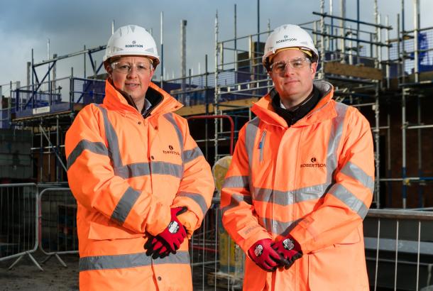 Two people standing in front of a Robertson Construction Yorkshire & East Midlands construction site, wearing personal protective equipment