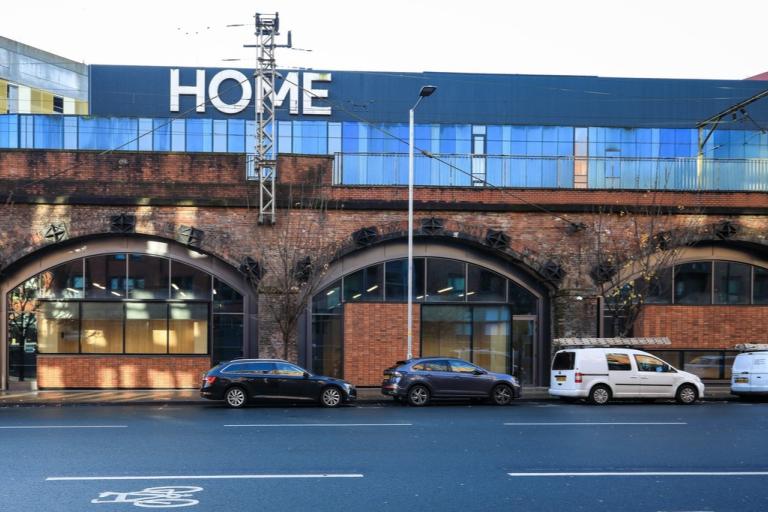 Street view of the new HOME Arches artistic community hub in Manchester, displaying a red-brick and glass facade below an existing railway