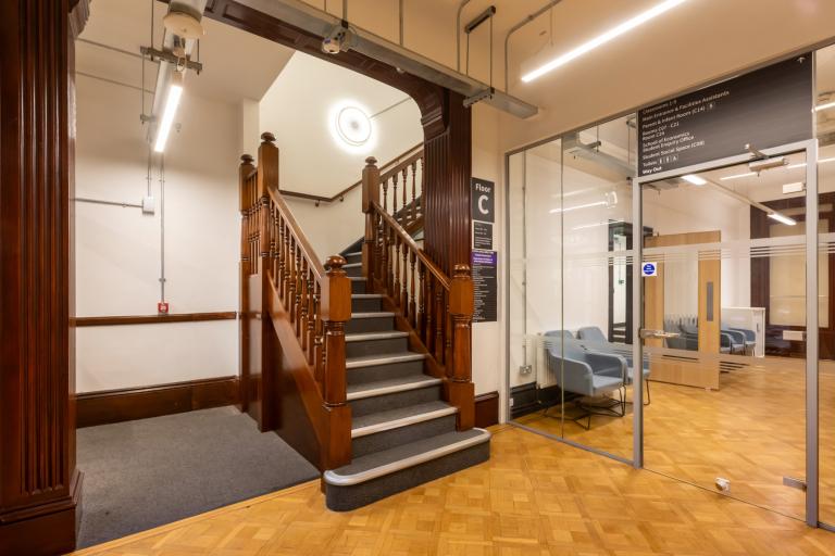 University building's interior showing a hardwood staircase, well-lit space and glass doorway leading to classrooms