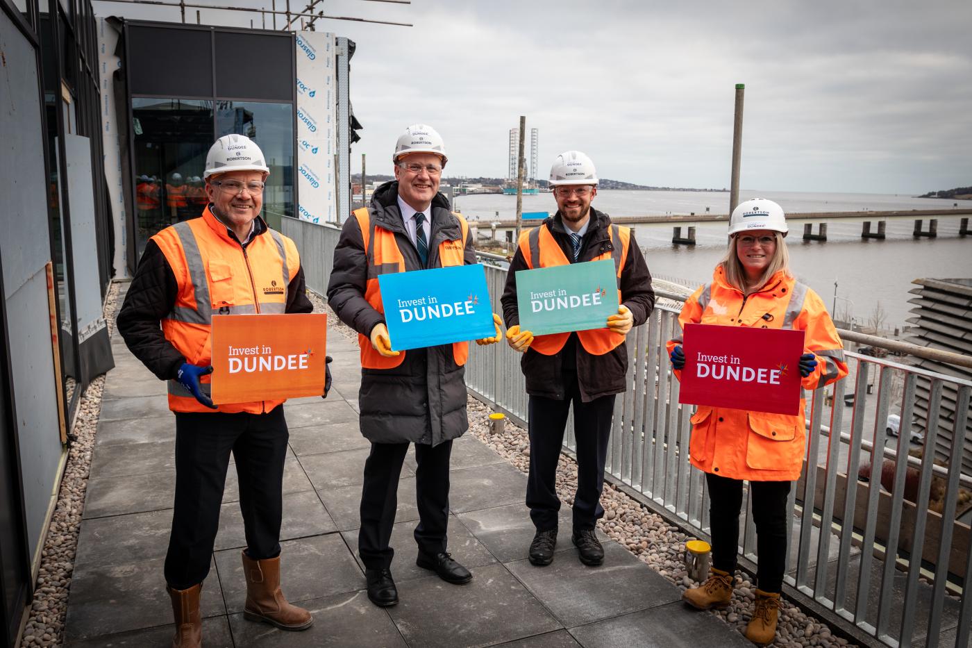 Four people at the construction site of James Thomson House, being delivered by Robertson Construction, holding signs reading 'Invest In Dundee'