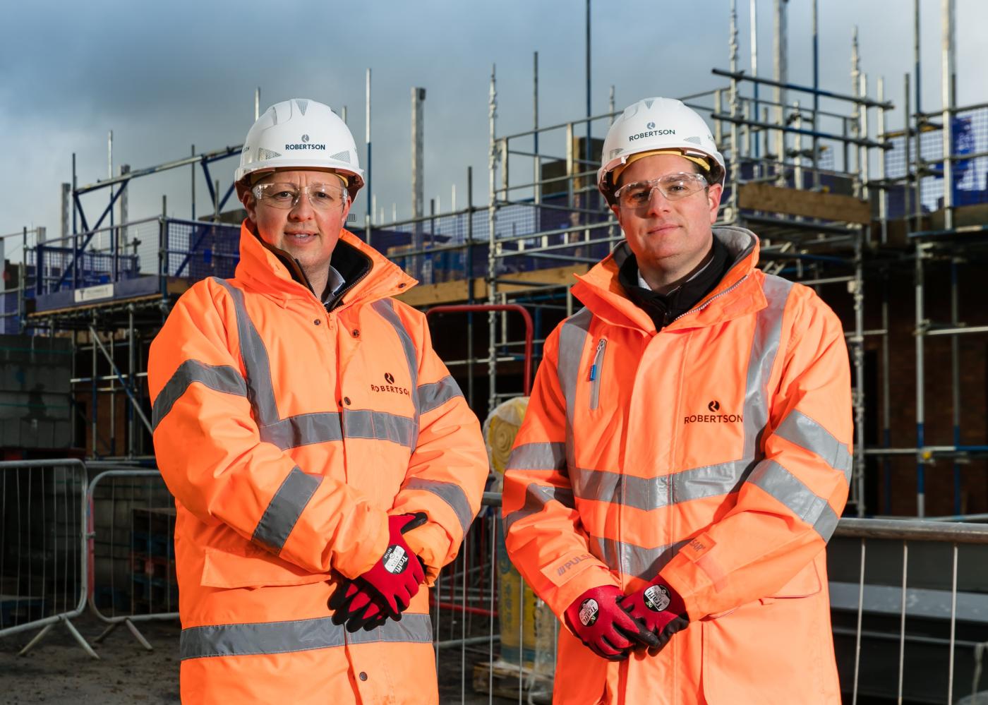 Two people standing in front of a Robertson Construction Yorkshire & East Midlands construction site, wearing personal protective equipment