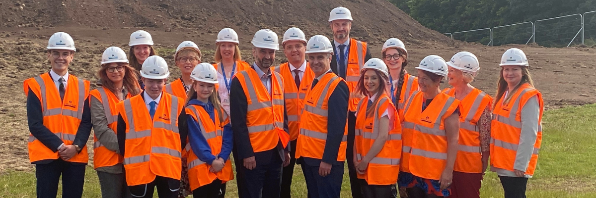 A group of workers and children standing in a row on a construction site wearing white hard hats and orange vests